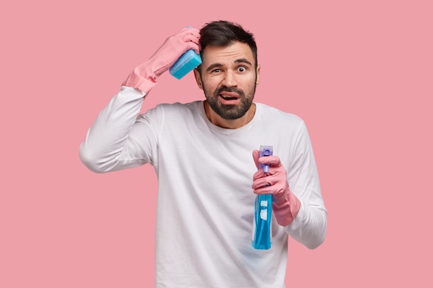 Housekeeping concept. Frustrated bearded young guy scratches head in bewilderment, has upset facial expression, wears casual white sweater