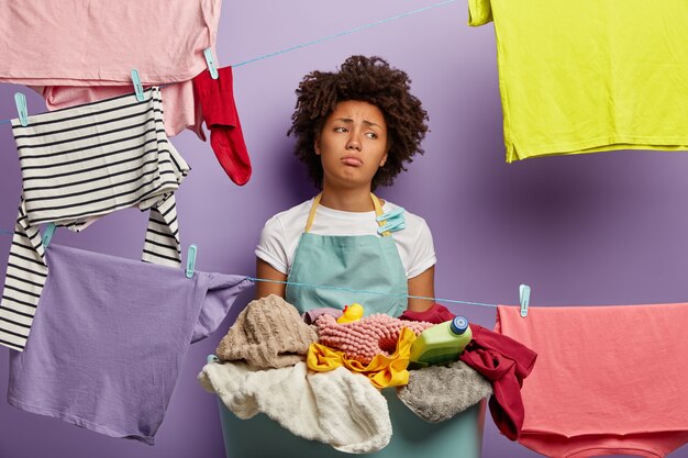 Housekeeping and washing concept. Dissatisfied sad young woman has Afro hairstyle, hangs clothes on clothes lines with clips, does laundry at home