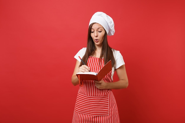 붉은 벽 배경에 격리된 줄무늬 앞치마 흰색 티셔츠 토크 셰프 모자를 쓴 주부 여성 요리사 또는 제빵사. 웃는 여자는 메모장 요리 책과 펜을 들고 있습니다. 복사 공간 개념을 비웃습니다.