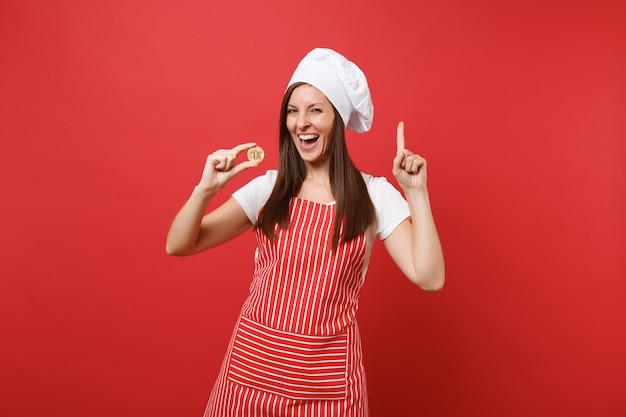 붉은 벽 배경에 격리된 줄무늬 앞치마 흰색 티셔츠 토크 셰프 모자를 쓴 주부 여성 요리사 또는 제빵사. 비트코인, 비트코인 미래 화폐를 들고 웃는 여자. 복사 공간 개념을 비웃습니다. 프리미엄 사진