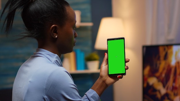 Housewife holding smartphone with chroma screen on hand looking at mockup. Reading on green screen template chroma key isolated mobile phone display using techology internet sitting on cozy couch