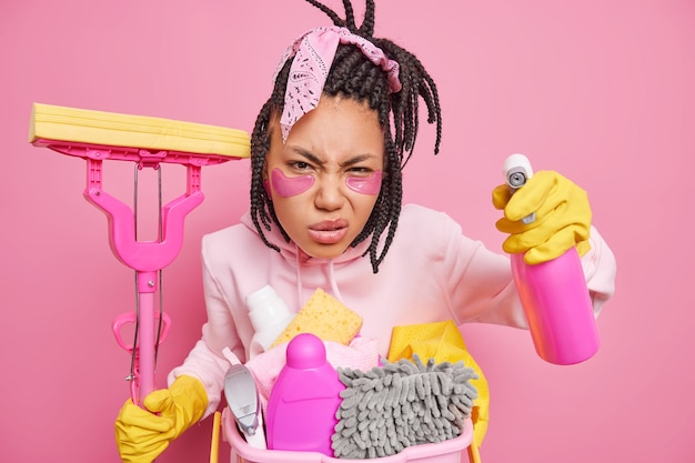 主婦は掃除道具を持ってポーズをとる 純度を気にする にやにや笑う 顔は綿密に何か汚いものを見つめる ピンクに隔離されたモップ洗浄用洗剤を持つ