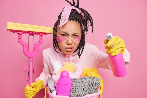 housewife poses with housecleaner tools cares about purity smirks face looks scrupulously at something dirty holds mop cleaning detergent isolated on pink 