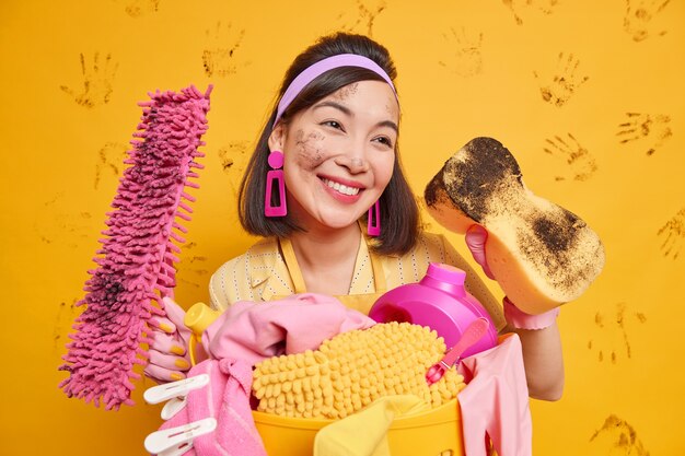 housewife remains beautiful even during house cleaning has happy dreamy expression wears headband earrings poses with dirty sponge mop stands near basket full of laundry isolated on yellow