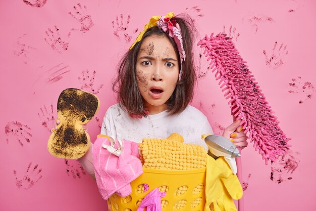 housewife stares with widely opened mouth shocked to have much housework holds cleaning tools tidies up room stands near laundry basket on pink 