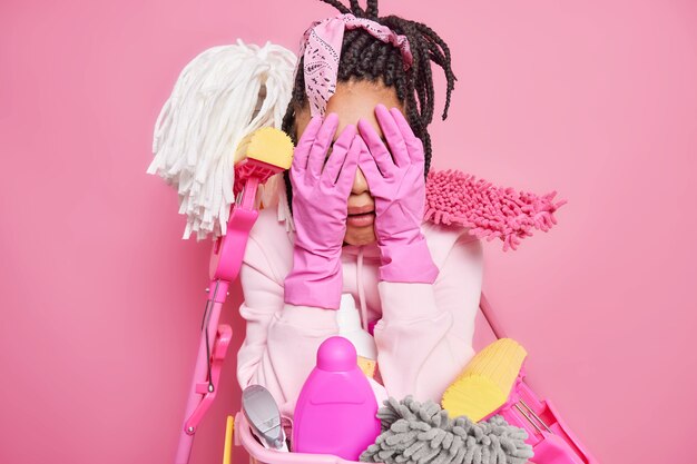 housewife tired of cleaning up all house cries and cons face with hands in rubber gloves surrounded by cleaning supplies expresses negative emotions isolated on pink 