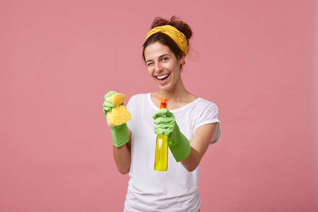 Housewife in white t-shirt and green gloves holding sponge and cleaner in hands blinking with her eyes having glad expression while washing. young pretty female doing domestic work