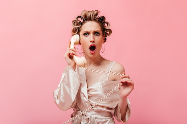 Free photo housewife with curly hair dressed in silk robe looks indignantly and shocked at front and speaks on phone
