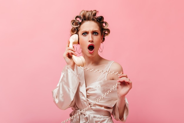 Housewife with curly hair dressed in silk robe looks indignantly and shocked at front and speaks on phone