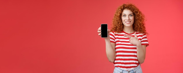 無料写真 このアプリはどうですか赤毛の巻き毛のフレンドリーなかわいい女の子は、友人がスマートフを保持するオンラインストアを見つけるのを助けます