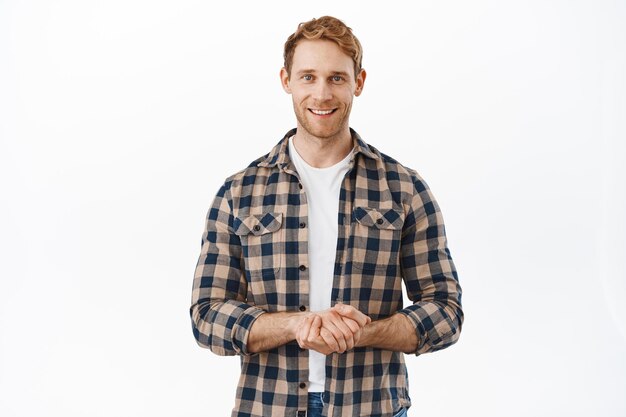 How may I assist you. Smiling pleasant man with red hair, looking content and determined, holding hands together, ready to offer his help, assisting clients, standing over white background