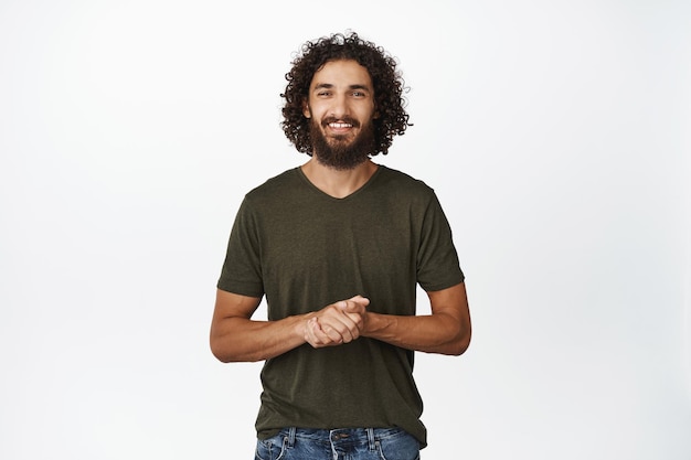 Come posso aiutarti sorridente giovane ragazzo moderno con la barba in attesa e guardando fiducioso che assiste in piedi su sfondo bianco