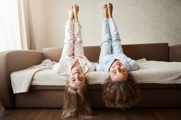 How nice to lie and fool around all day with girlfriend. Charming childish european women lying on sofa in nightwear with lifted hands and head upside down, hair touching floor, smiling broadly