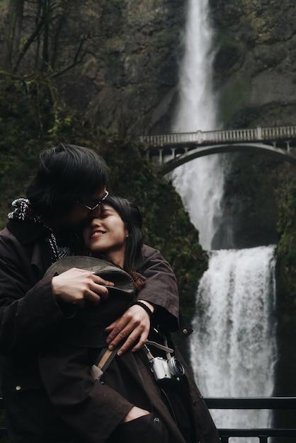 Free photo hugging asian couple stands before a beautiful waterfall in the mountains