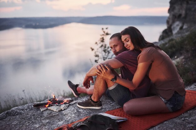 Hugging couple with backpack sitting near the fire on top of mountain enjoying view coast a river or lake.