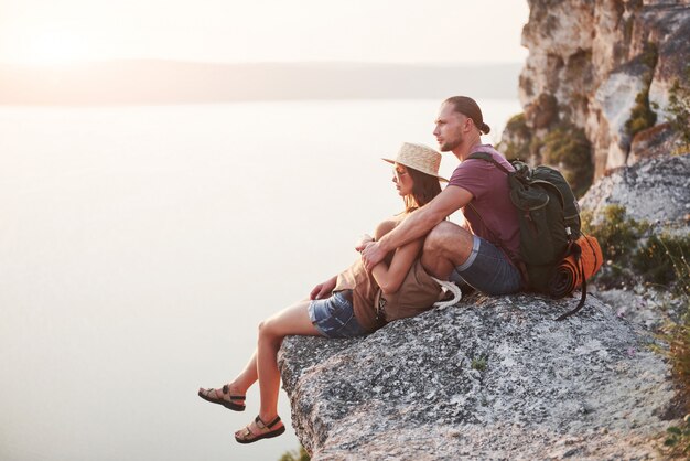 Hugging couple with backpack sitting on top of rock mountain enjoying view coast a river or lake.