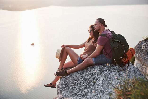 Обнимающ пар при рюкзак сидя на верхней части горы утеса наслаждаясь побережьем взгляда река или озеро.