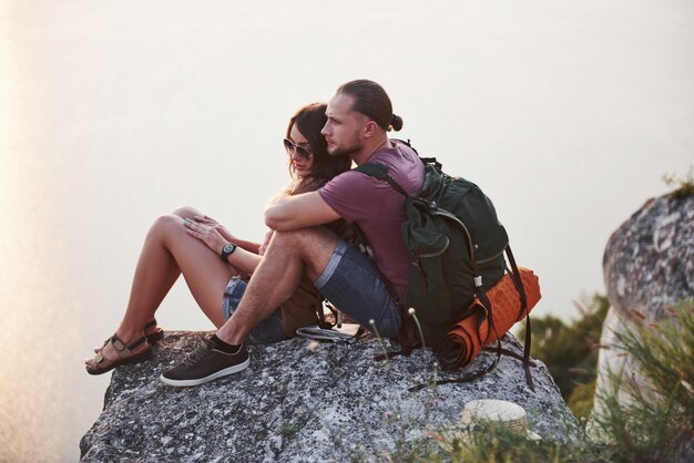川や湖の海岸を眺めながら岩山の上に座っているバックパックとカップルを抱き締めます。