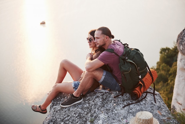 Hugging couple with backpack sitting on top of rock mountain enjoying view coast a river or lake.