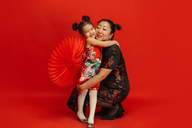 抱きしめて、幸せな笑顔。ハッピーチャイニーズニューイヤー2020。伝統的な服の赤い背景に分離されたアジアの母と娘の肖像画。お祝い、人間の感情、休日。コピースペース。