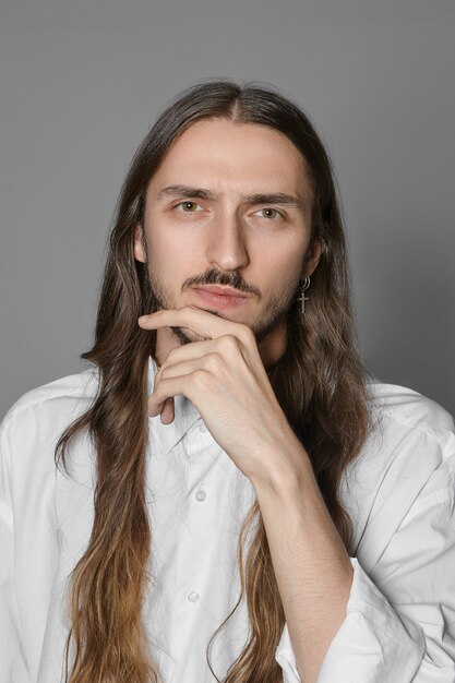 Human emotions and body language. Vertical picture of stylish extraordinary male with long brown hair and mustache keeping hand on his chin