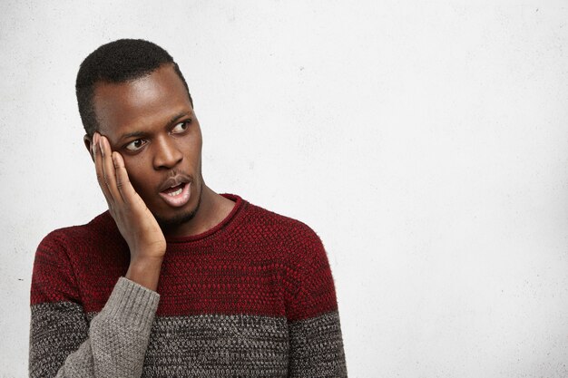 Human emotions and feelings. Handsome shocked African American male wearing casual sweater holding hand on cheek in surprise and astohisnment, looking at blank white copy space wall, mouth wide open