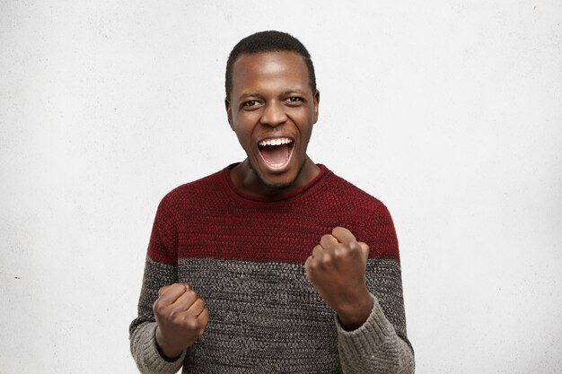 Human emotions and feelings. Happy lucky excited young dark-skinned male winner exclaiming, rejoicing at his success at work, saying yes, clenching fists while achieving his life and career goals