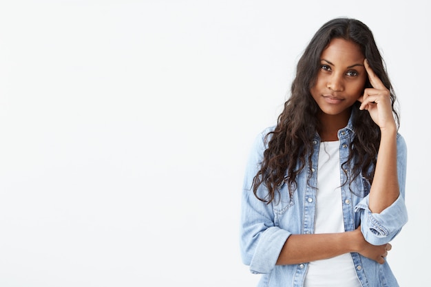 Human emotions, feelings, reaction and attitude. Attractive young dark-skinned woman in denim with long wavy hair squeezing eyes and keeping hand on temple in dissatisfaction and suspicion, feeling sc