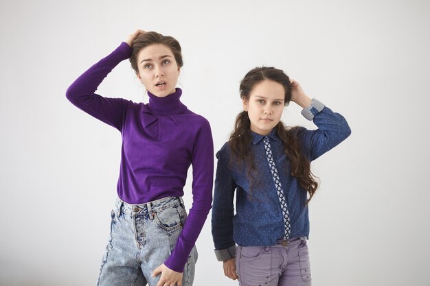 Human emotions, feelings, reaction and attitude. Isolated studio shot of two young sisters in stylish clothes standing at white wall