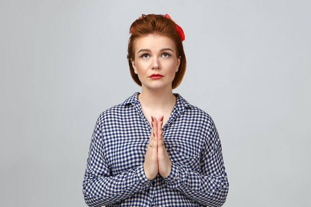 Human emotions, feelings and reaction. Stylish young European female in checkered shirt looking up with mournful hopeful facial expression, keeping hands in namaste, praying. Please forgive me