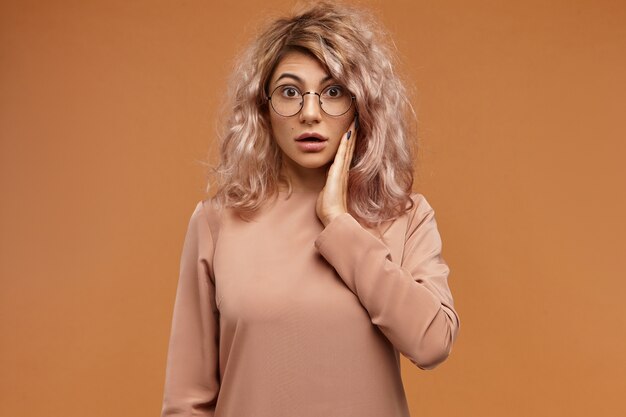 Human emotions, reaction and feelings.  shot of emotional funny hipster girl in round eyeglasses, having puzzled look, holding cheek and opening mouth