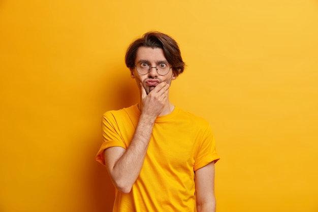 Human face expressions concept. Handsome adult European man holds chin pouts lips makes funny grimace wears round transparent glasses and casual t shirt isolated over yellow  wall.