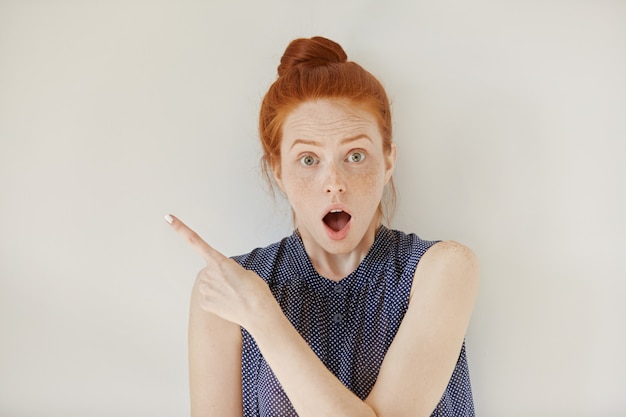 Human face expressions, emotions and feelings. Astonished and shocked young redhead female in sleeveless shirt pointing at blank wall