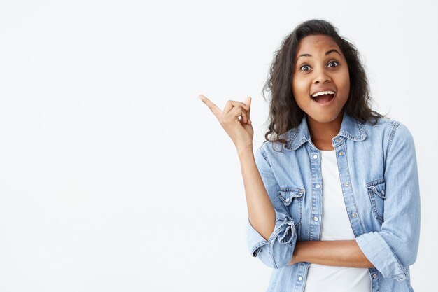 Human face expressions, emotions and feelings. Astonished and surprised young Afro-american female in light-blue denim shirt pointing at blank wall, surprised with sale prices, keeping her mouth