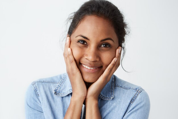 Человеческое выражение лица и эмоции. Портрет молодой афроамериканской женщины в голубой джинсовой рубашке, смотрит счастливой, держа голову обеими руками, с улыбкой.