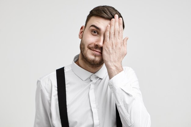 Human facial expressions and body language. Isolated shot of positive young bearded businessman covering one half of his face and smiling joyfully at camera. Optics, vision, eyesight and ophthalmology