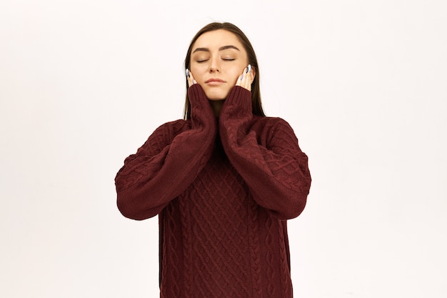 Human facial expressions and body language. Studio image of attractive stylish young European female closing eyes, holding hands on her cheeks, dreaming, meditating or having nap, wearing jumper