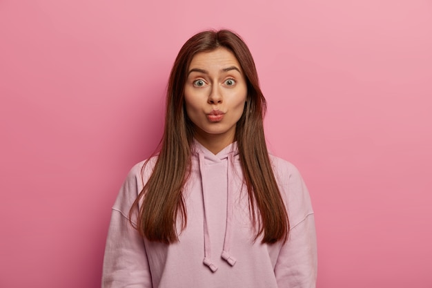 Human facial expressions concept. Beautiful teenager keeps lips rounded, has healthy skin, going to kiss someone, wears pastel rosy hoodie