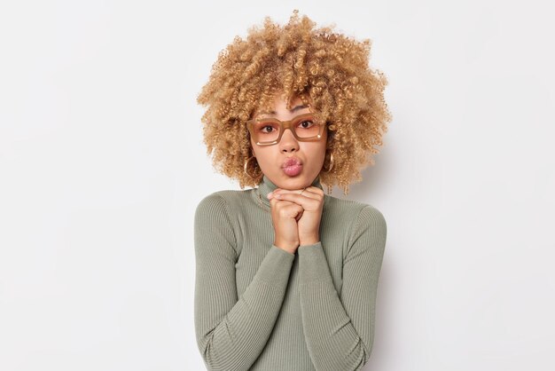 Human facial expressions concept Pretty curly haired woman keeps lips folded hands under chin wears spectacles and turtleneck isolated over white background Wondered female model blows mwah