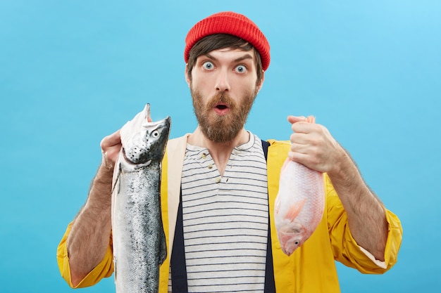 Бесплатное фото Выражения лица, эмоции и чувства человека. забавный изумленный молодой рыбак в красной шляпе и желтом плаще позирует у стены с двумя рыбками, удивленный прекрасным уловом