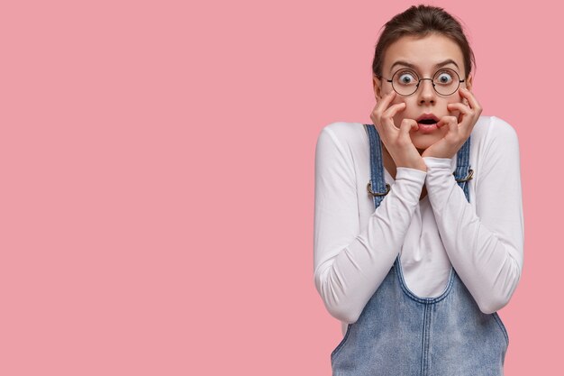 Human facial expressions and emotions concept. Woman in despair and shock, stands in stupor against pink background, touches face