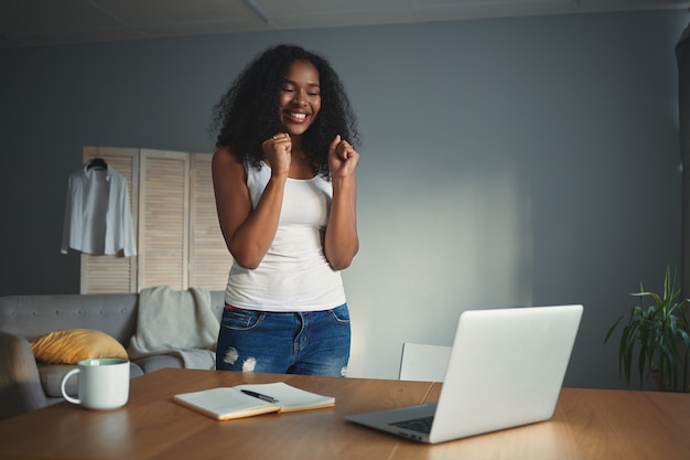 Human facial expressions and emotions. Euphoric emotional young mixed race female clenching fists, being excited with good news, success or promotion at work, standing at desk with laptop and diary