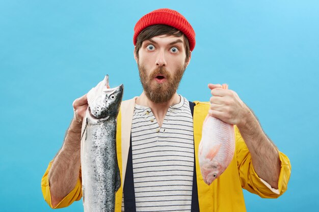 Human facial expressions, emotions and feelings. Funny astonished young fisherman wearing red hat and yellow raincoat posing against wall with two fish, surprised with fine catch