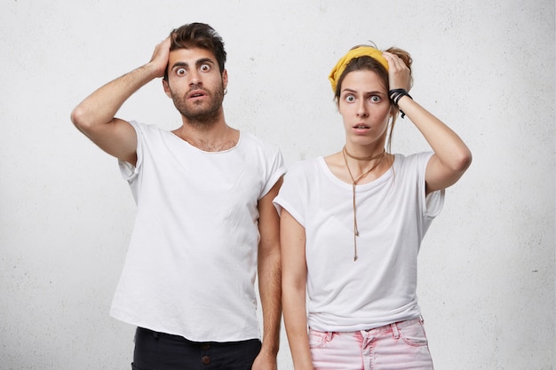 Human facial expressions, emotions, feelings, reaction and attitude. Indoor shot of shocked forgetful young Caucasian man and woman holding hands on heads and looking with shock and terror