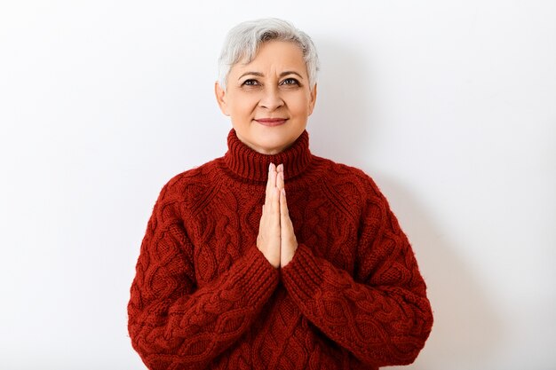 Human facial expressions, emotions, feelings and reaction. Isolated image of positive cheerful retired woman with short hair looking up with happy smile, holding hands in prayer, having hopeful look