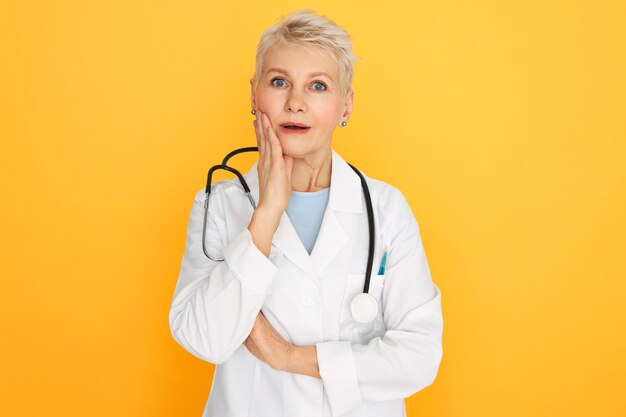 Human facial expressions, emotions and feelings. Studio image of emotional surprised retired woman practitioner holding hand on cheek and opening mouth, shocked with anamnesis or diagnosis