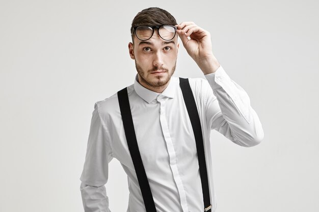 Human facial expressions and emotions. Portrait of emotional fashionable young bearded Caucasian male dressed in formal wear lifting his stylish eyeglasses, having confused questioning look