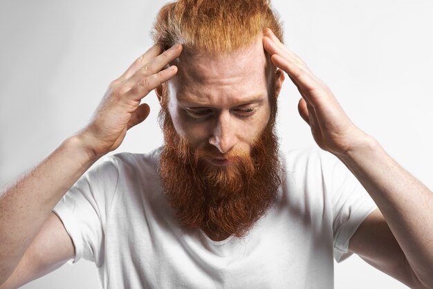 Human facial expressions, emotions, reaction and attitude. Portrait of frustrated young Caucasian bearded male feeling stressed, having some problem, looking down, holding hands on his head