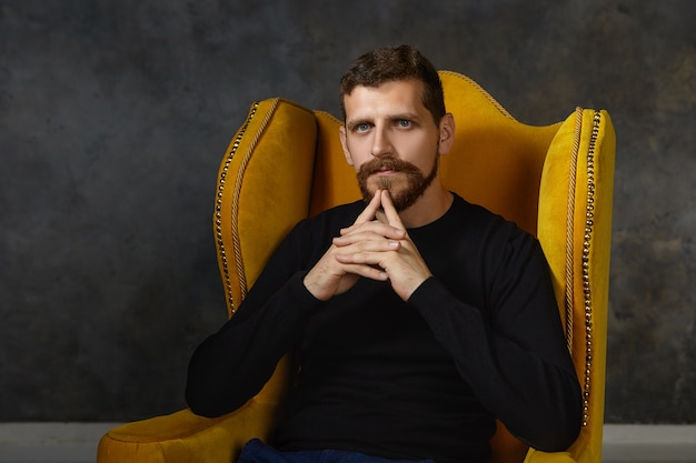 Human facial expressions, feelings and body language. Serious fashionable young bearded businessman touching chin, having pensive look while thinking over business plan and ideas in armchair