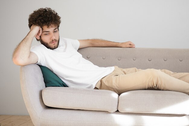 Human facial expressions, feelings and emotions. Horizontal portrait of handsome stylish unshaven male in white t-shirt and beige jeans feeling bored and lonely, spending time at home alone